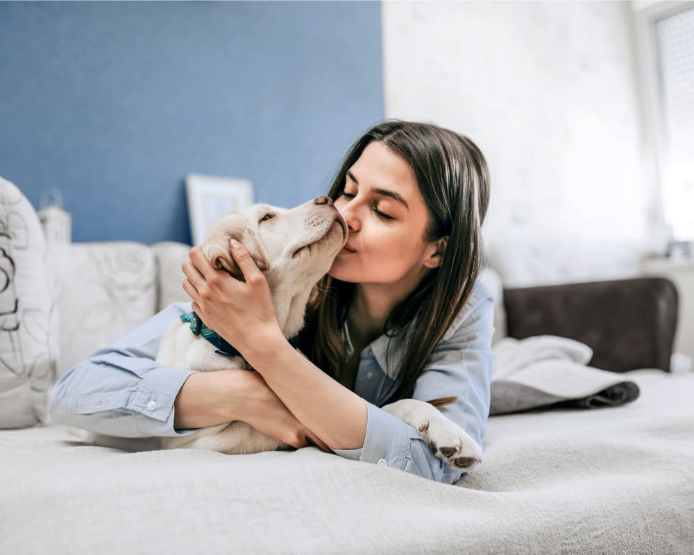 A woman lays on a bed with a young blonde dog and kisses its snout.